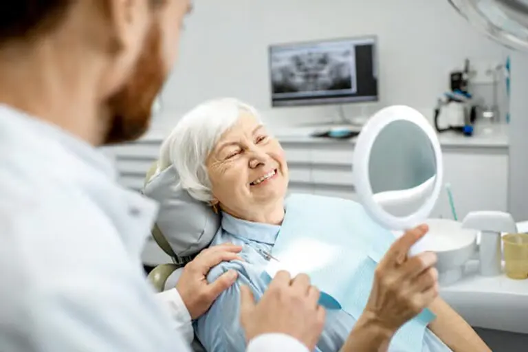 Elderly woman in the dentist chair smiling at her reflection with new implant dentures